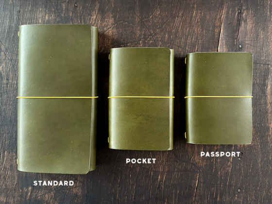 Tri-fold traveler journal leather covers in three sizes (standard, pocket, and passport) with elastic bands on a wooden table, labeled by size.