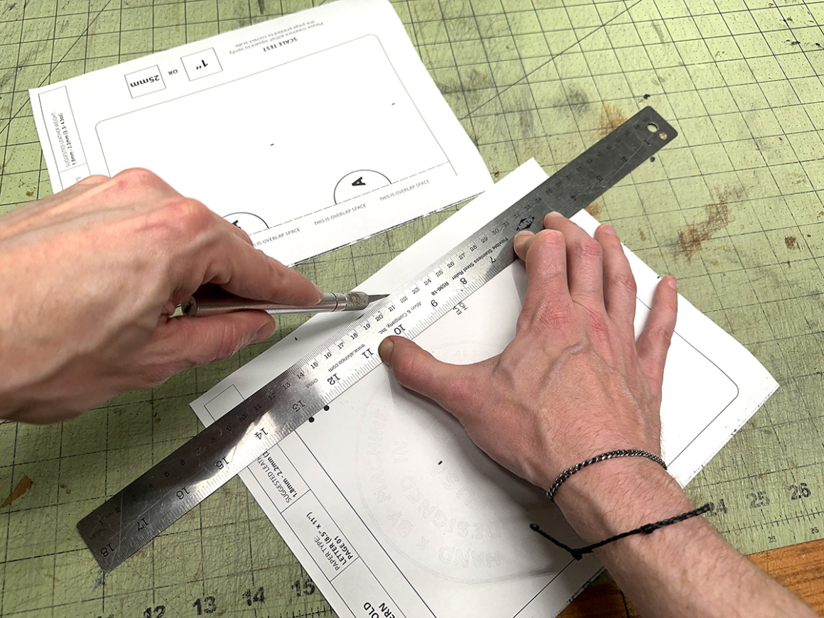 Trimming the A5 bifold leather notebook cover pattern page along a straight edge ruler with an X-Acto knife on a cutting mat
