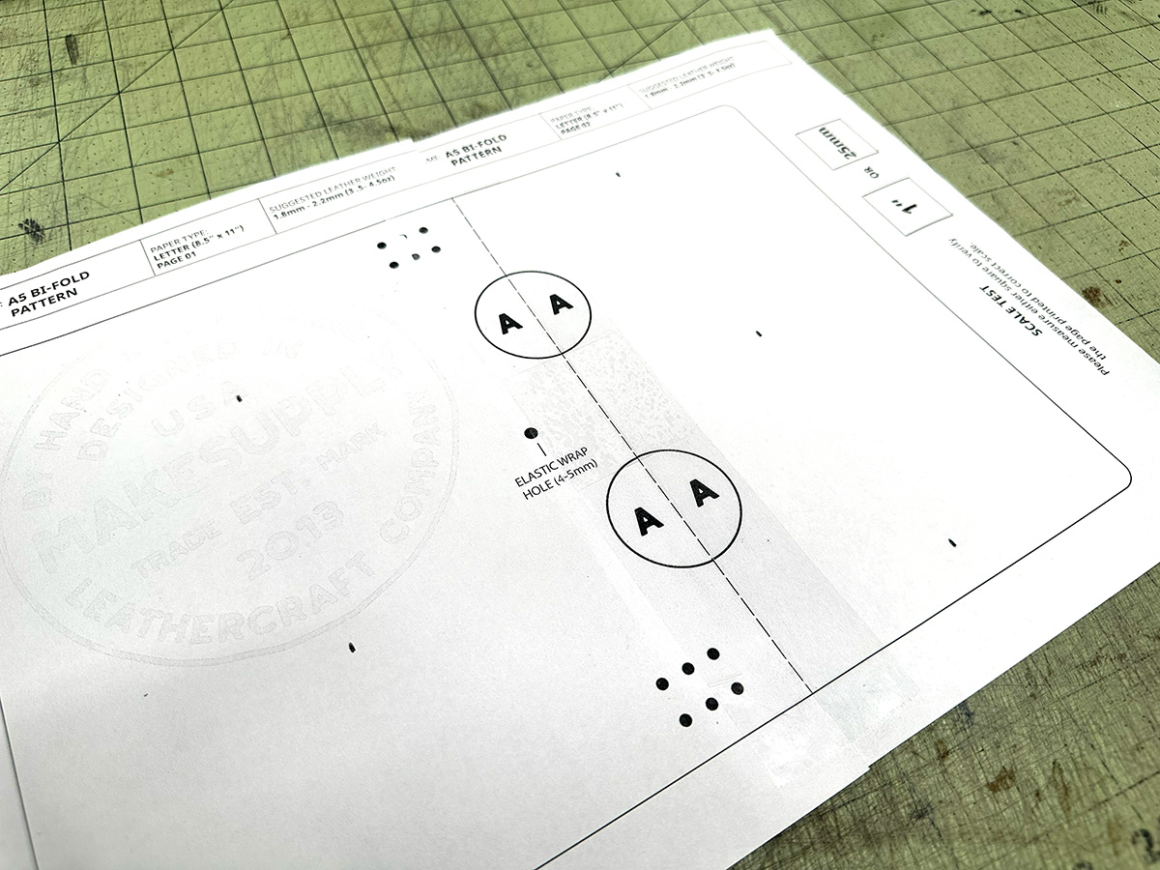 `Two pages of the A5 bifold leather notebook cover pattern assembled and aligned on a cutting mat showing the elastic wrap hole placement and registration marks`
