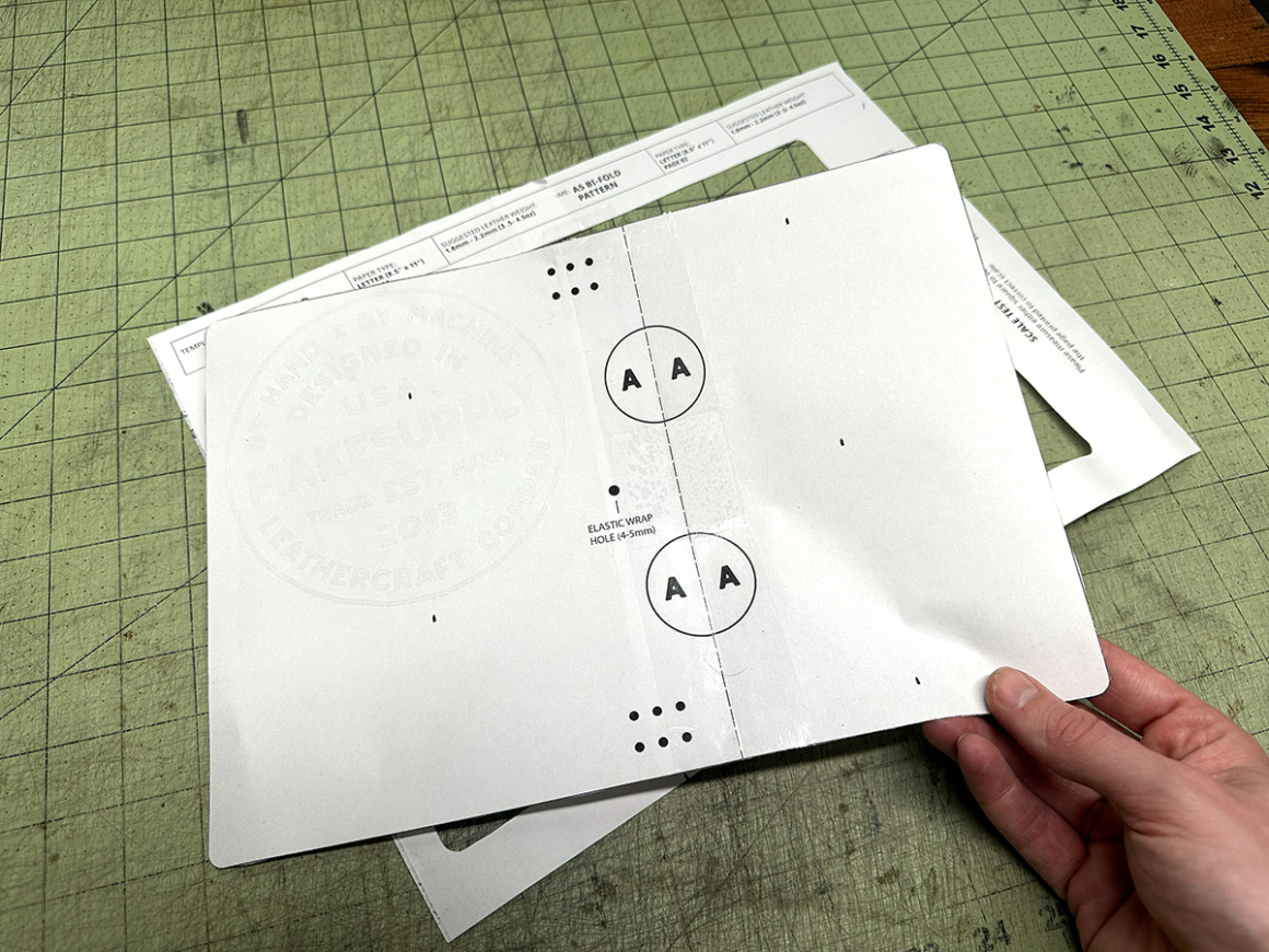 `Assembled A5 bifold leather notebook cover pattern pages taped together and held over a cutting mat showing the full cover layout`